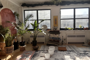 Studio space lit by sunlight, meditation cushions and singing bowls visible, along with plants